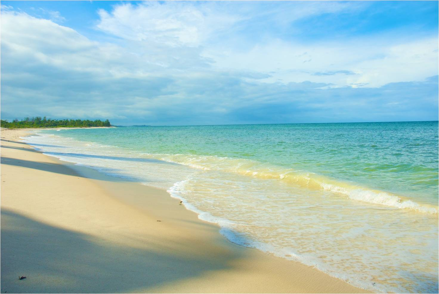 Gentle turquoise waves rolling onto a sandy beach under a blue sky with clouds